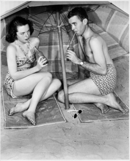 Young Couple Sitting Under Beach Umbrella Drinking Poster Print - 18 x 24 in.