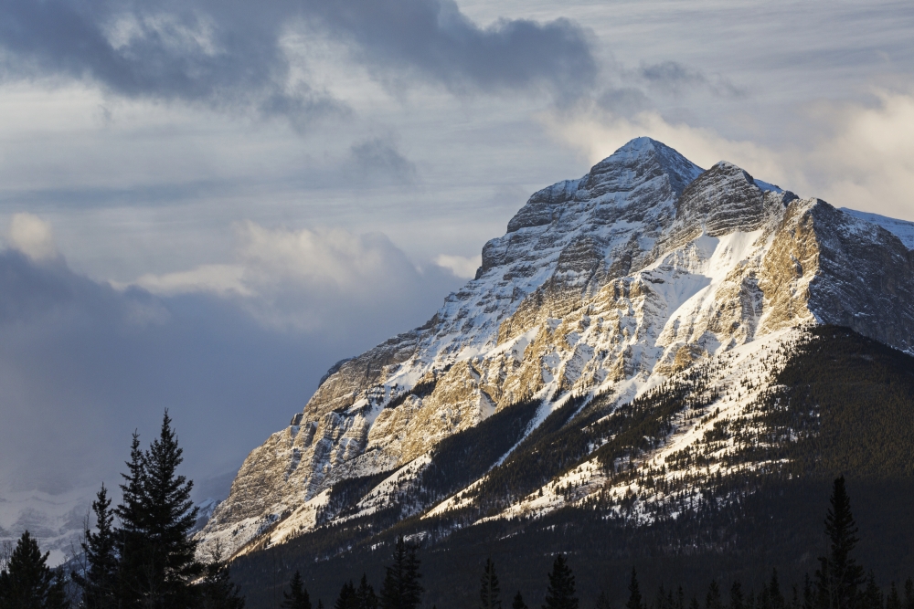 Sun Breaking Through The Clouds Partially Lighting A Snow Covered Mountain with Clouds in The Sky - Kananaskis Country A Poster Print - 19 x 12 in.