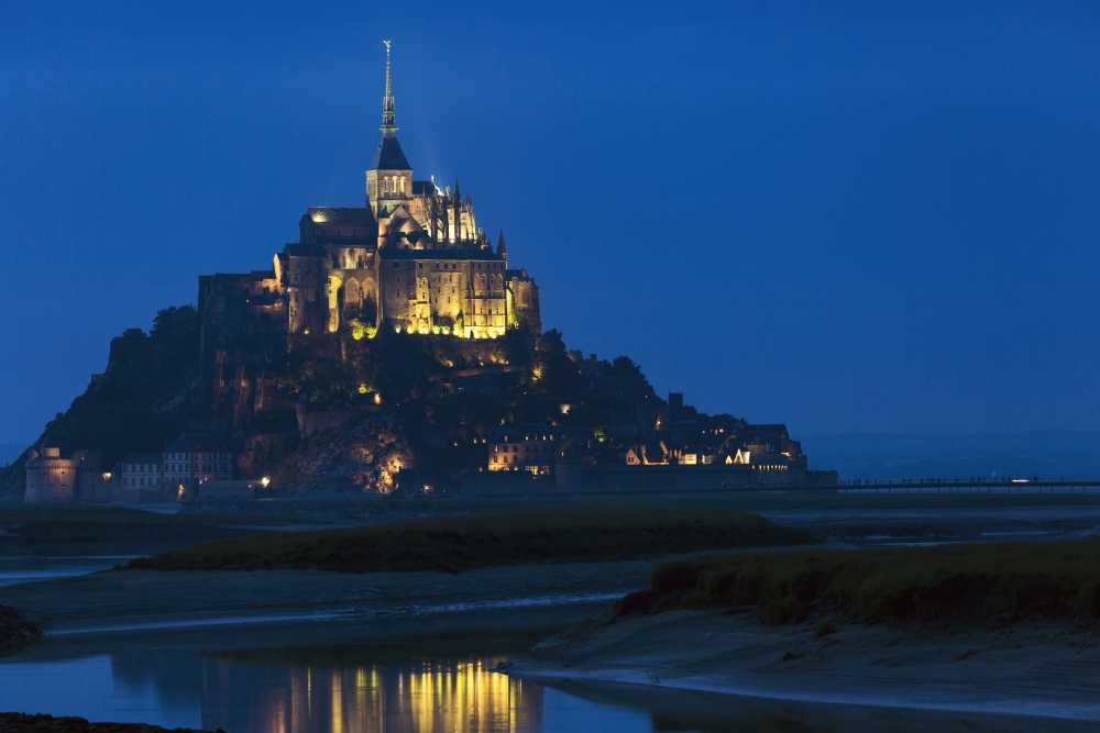 Night Image of Large Stone Abbey Perched on Rocky Island at Low Tide Lit with Flood Lights with Reflection in Water Poster Print - 19 x 12 in.